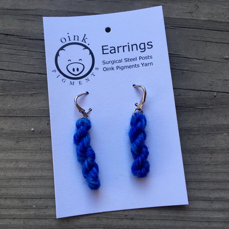 miniature skeins of yarn dangle from steel earring hooks on a white card with the Oink Pigments logo and information at the top.