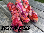 three brightly colored skeins of coral, red, and orange yarn sit on a wooden background. Black text at the bottom left reads "hot mess"