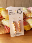 miniature skein earrings rest on a white card with the Oink Pigments logo and text at the top of the card. The card rests on a stacked pair of skeins of yarn. 