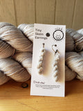 miniature skeins of yarn dangle from steel earring hooks on a white card with the Oink Pigments logo and information at the top.  The card is resting against two skeins of yarn.