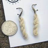 miniature skeins of yarn dangle from steel earring hooks on a white card with the Oink Pigments logo and information at the top. A quarter rests to the left of frame for size reference. 