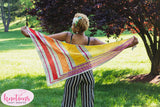 A brightly colored shawl is held behind a medium skinned person standing in a green field.  There is a dark reddish tree in the right side of the frame and a small pink stamp in the bottom left that reads Knotions, Craft Smarter.
