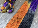 A bold swatch of speckled orange, deep black and rich purple yarn rests beside a stack of three skeins in the upper left corner.  The background is a light grey wood. 