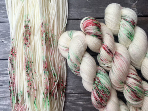 An unfurled skein rests on a dark wooden background beside three twisted up skeins.  The yarn is white with red and green speckles. 