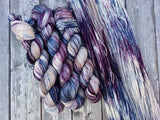 Three skeins lie in twists to the left, beside a swath of yarn resting against a grey wooden background. 