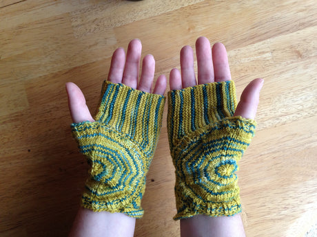 light skinned hands, clad in a stripey, swirley fingerless mitts hover above a light wooden background.