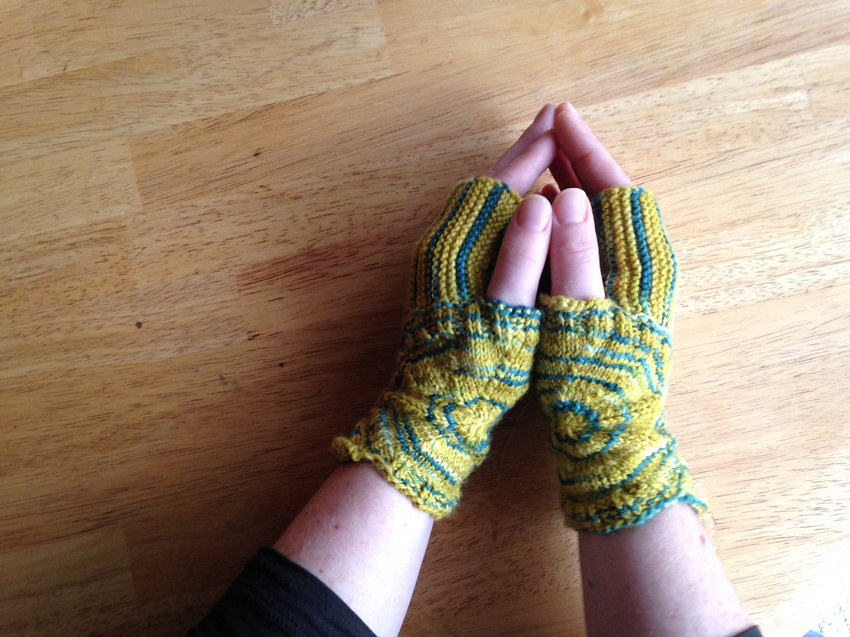a pair of light skinned hands rest on a light wooden background. Blue and yellow fingerless mitts wrap around the wrists and extend halfway up the fingers. 