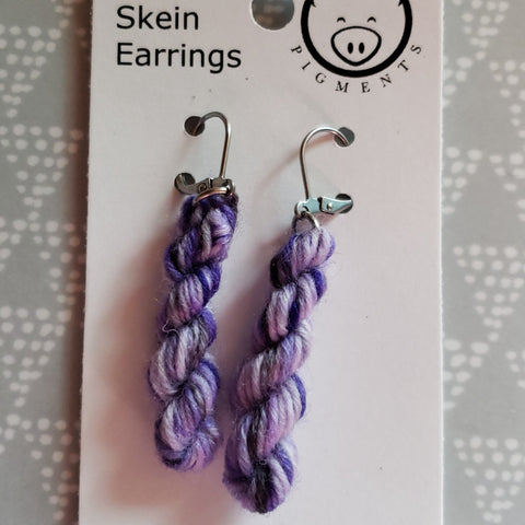 Miniature skeins dangle from steel earring hooks on a white card with the Oink Pigments logo at the top.  A grey background with spotty white triangles can be seen on either side of the card.