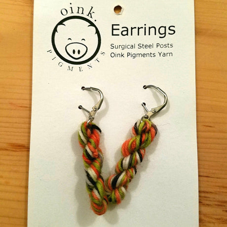 miniature skeins of yarn dangle from steel earring hooks on a white card with the Oink Pigments logo and information at the top.