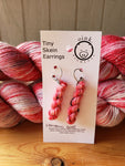 miniature skeins of yarn dangle from steel earring hooks on a white card with the Oink Pigments logo and information at the top. Two skeins of yarn rest in the background.
