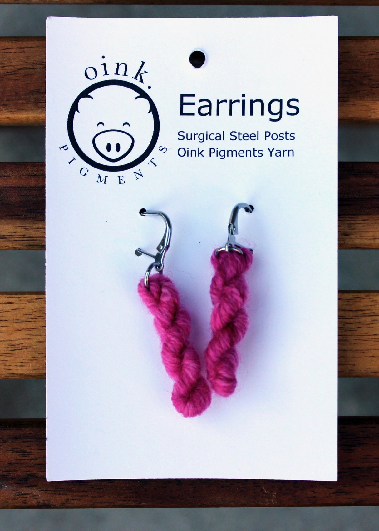 miniature skeins of yarn dangle from steel earring hooks on a white card with the Oink Pigments logo and information at the top.