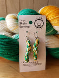 miniature skeins of yarn dangle from steel earring hooks on a white card with the Oink Pigments logo and information at the top. Two full skeins sit in the background with the earring card resting against them.