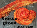 skein of yarn twisted in a spiral and one laid out both are a bright orange with splashes of deep orange and black speckles, black text reads "Spiked cider July 20202"