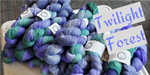 a delightful pile of twisted skeins of yarn, in various bases rests atop a white table.  The yarn is colored in tones of periwinkle, lilac, purple, and a soft teal-green. Lilac text on light grey background on the right reads "twilight forest"